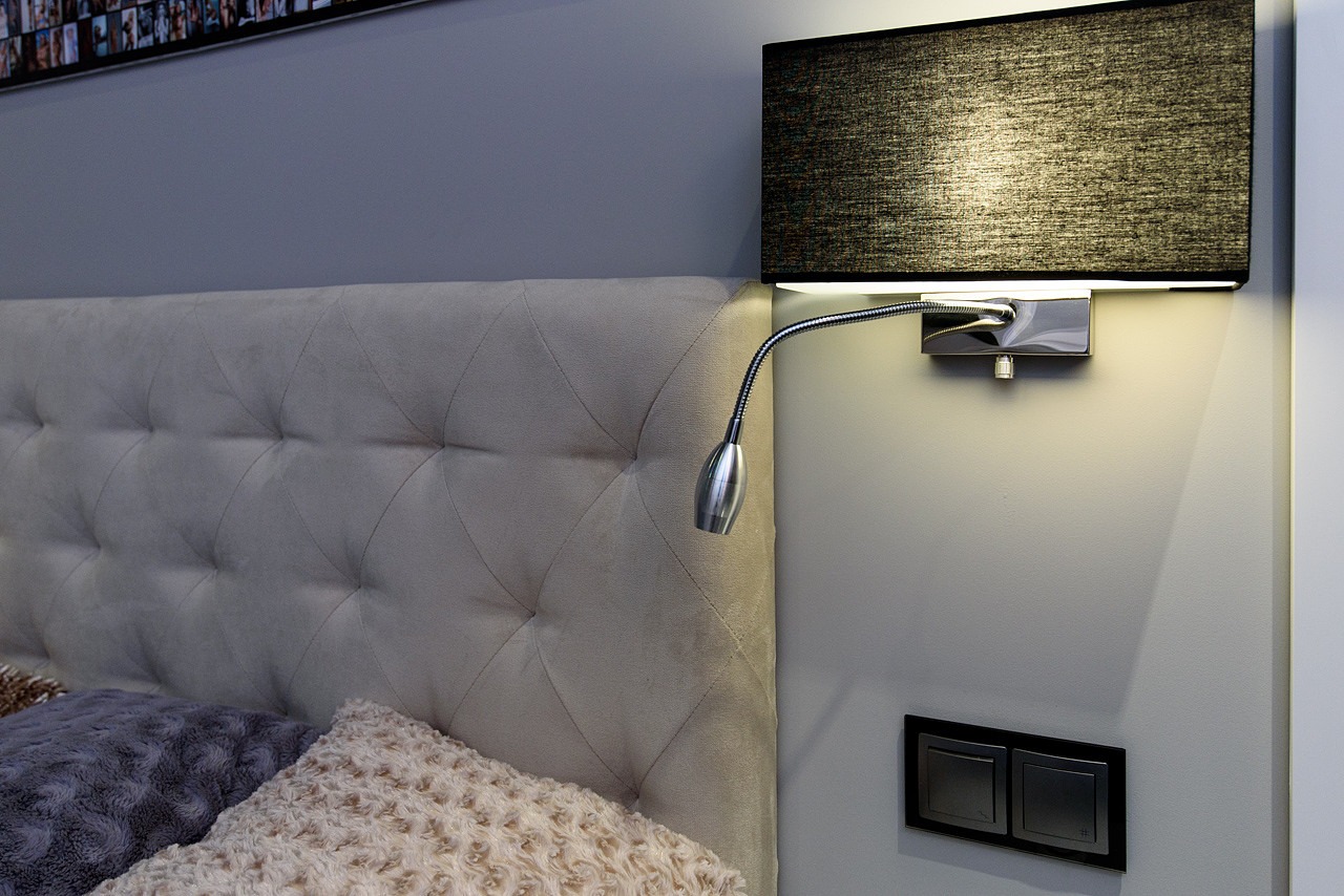 Tapicerowane wezgłowie łóżka w kolorze beżowym, z pikowaniem w romby, oświetlone regulowaną lampką nocną z czarnym abażurem i metalowym ramieniem, obok włącznik światła.