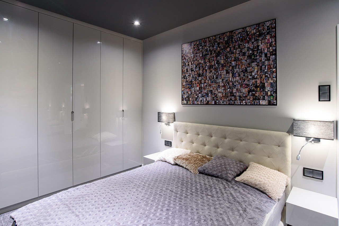 Nowoczesna sypialnia z dużym łóżkiem, błyszczącą szafą w zabudowie, fotomozaiką na ścianie i lampkami nocnymi z abażurami.