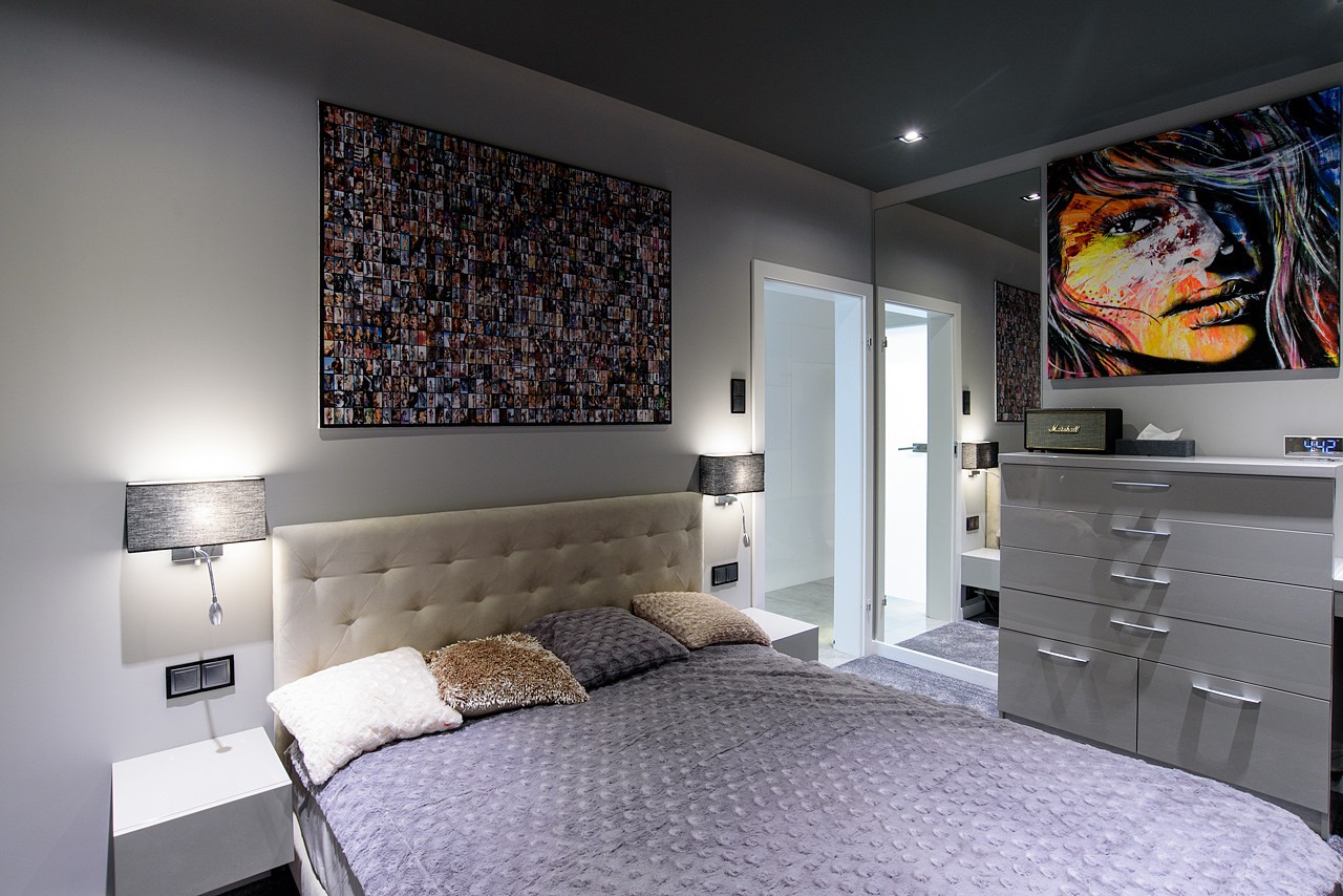 Nowoczesna sypialnia w szarościach z łóżkiem z pikowanym zagłówkiem, lampkami nocnymi z abażurami, obrazem z mozaiką zdjęć i portretem kobiety, oraz lustrzaną szafą.
