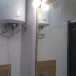 Biały bojler elektryczny zamontowany na kafelkowanej ścianie w łazience, widoczne podłączenia wodne i elektryczne, odbicie bojlera w lustrze z drewnianą ramą.