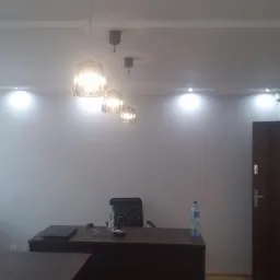 Białe biuro z trzema wiszącymi lampami z kloszami, biurkiem z laptopem i butelkami wody, oknem z żaluzjami poziomymi oraz zamykanymi drzwiami wejściowymi