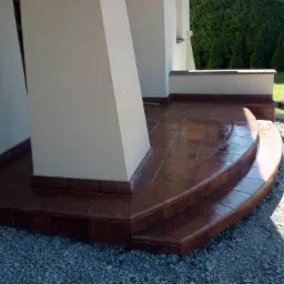 Zewnętrzne schody wejściowe wyłożone brązowymi płytkami ceramicznymi, z widocznymi fugami i zaokrąglonymi krawędziami, prowadzące na taras, z kolumnami podtrzymującymi zadaszenie i żwirowym podłożem.