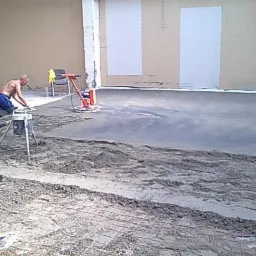 Pracownik w trakcie wygładzania wylewki betonowej za pomocą zacieraczki mechanicznej; widoczne narzędzia i siatka zbrojeniowa w tle.