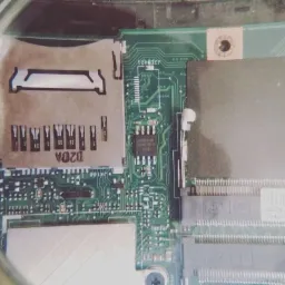 Powiększenie fragmentu zielonej płyty głównej laptopa z widocznym gniazdem SD, elementami elektronicznymi i ścieżkami, oglądane przez szkło powiększające.