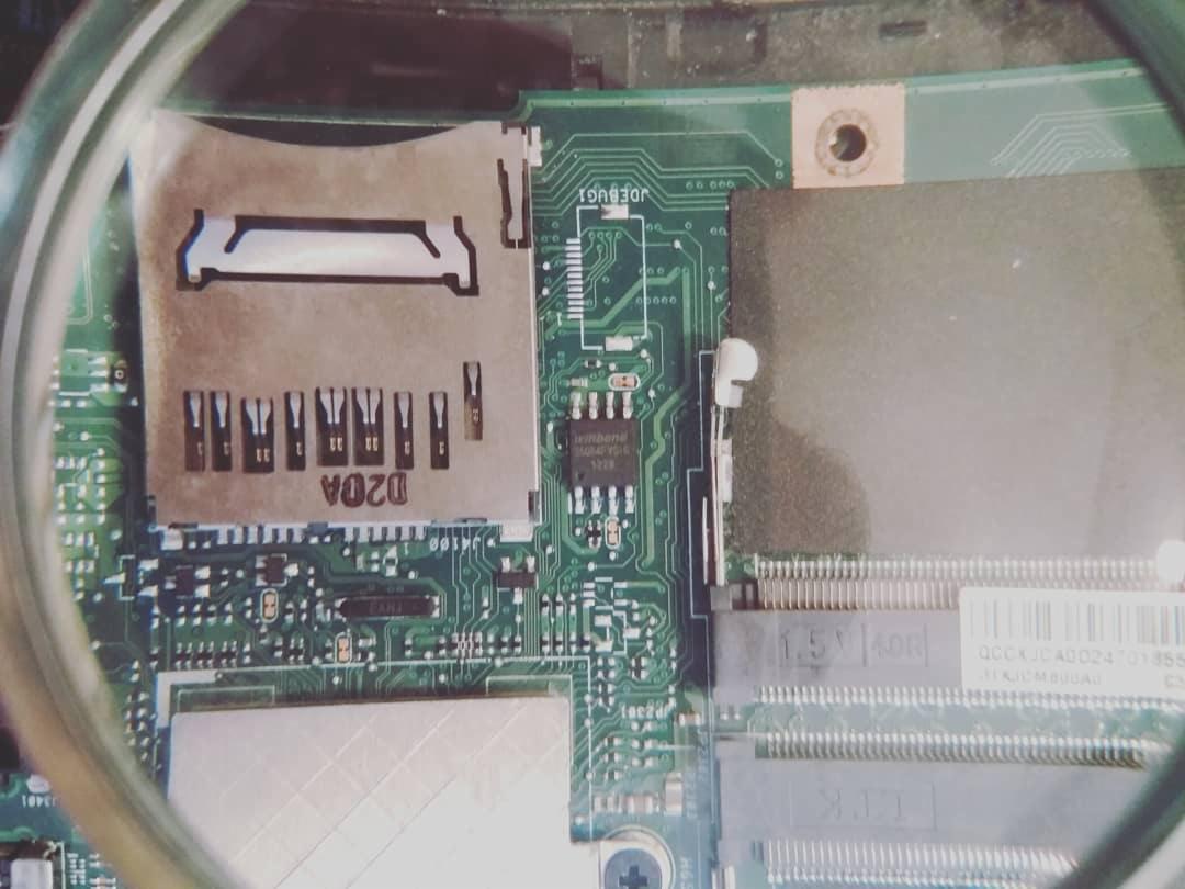 Powiększenie fragmentu zielonej płyty głównej laptopa z widocznym gniazdem SD, elementami elektronicznymi i ścieżkami, oglądane przez szkło powiększające.