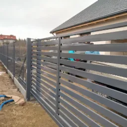 Nowoczesne, poziome przęsła w szarej, metalowej konstrukcji ogrodzenia posesji z widoczną siatką ogrodzeniową w tle i fragmentem domu z szarą dachówką.