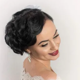 Uczesanie w stylu lat 20. z ciemnymi, falowanymi włosami, podkreślone oczy i ciemnoczerwone usta, widoczne długie, srebrne kolczyki i fragment koronkowej sukienki.