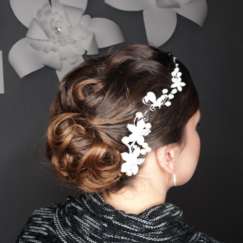 Upięcie ślubne z brązowych włosów z wplecioną ozdobną spinką z motywem kwiatowym i perłami, widok z tyłu na tle szarej ściany z papierowymi kwiatami.