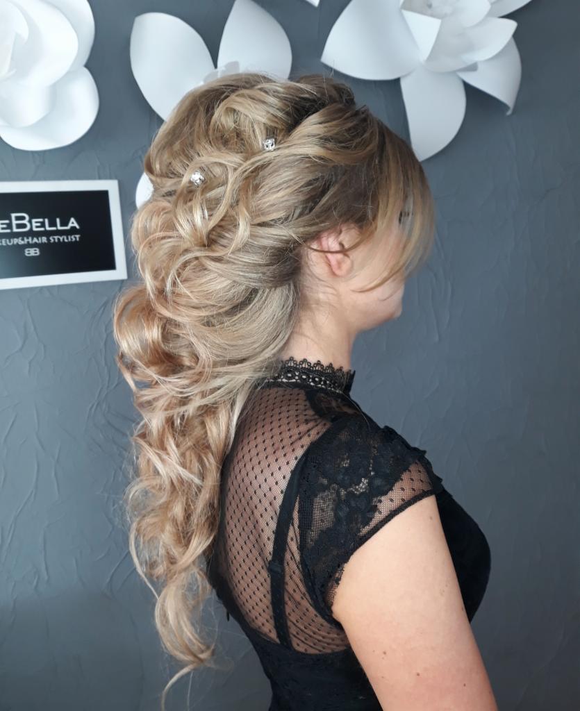 Elegancka upięta fryzura z blond pasemkami, ozdobiona błyszczącymi wsuwkami, na tle szarej ściany z papierowymi kwiatami i logo studia.