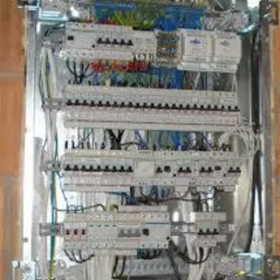 Rozdzielnica elektryczna w trakcie montażu, widoczne liczne zabezpieczenia, przewody o różnych kolorach i przekrojach, częściowo zamontowana w metalowej obudowie w surowym wnętrzu budynku.