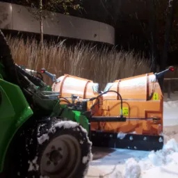 Zielony traktor z pomarańczowym pługiem do odśnieżania, pokryty śniegiem, na tle zaśnieżonej trawy i budynku w nocy.