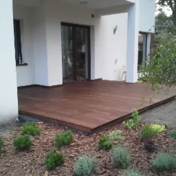 Nowoczesny taras z ciemnego drewna przylegający do jasnego budynku, otoczony rabatą z niskimi roślinami i korą.