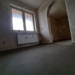 Świeżo wylana, szara posadzka w pomieszczeniu z oknem i grzejnikiem, widoczne ślady po pracach budowlanych na ścianach i łukowate przejście do kolejnego pokoju.