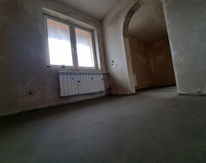 Świeżo wylana, szara posadzka w pomieszczeniu z oknem i grzejnikiem, widoczne ślady po pracach budowlanych na ścianach i łukowate przejście do kolejnego pokoju.