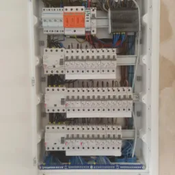 Otwarta biała skrzynka z zamontowanymi bezpiecznikami i przewodami na ścianie z surowym tynkiem.