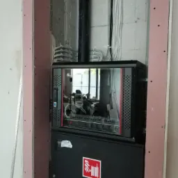 Szafa rack z oszklonymi drzwiami w surowym wnętrzu, widoczne kable i przewody, na dole szafka z naklejką ppoż.