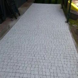 Jasnoszara, świeżo ułożona nawierzchnia z kostki brukowej przed domem, z widocznym fragmentem elewacji, złożonymi krzesłami i zielonym ogrodzeniem.