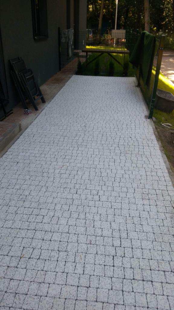 Jasnoszara, świeżo ułożona nawierzchnia z kostki brukowej przed domem, z widocznym fragmentem elewacji, złożonymi krzesłami i zielonym ogrodzeniem.