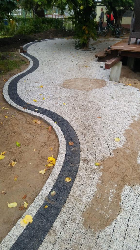 Kręta ścieżka z jasnoszarej kostki brukowej z ciemnoszarym pasem, prowadząca od trawnika do drewnianego tarasu, z widocznymi opadłymi żółtymi liśćmi.