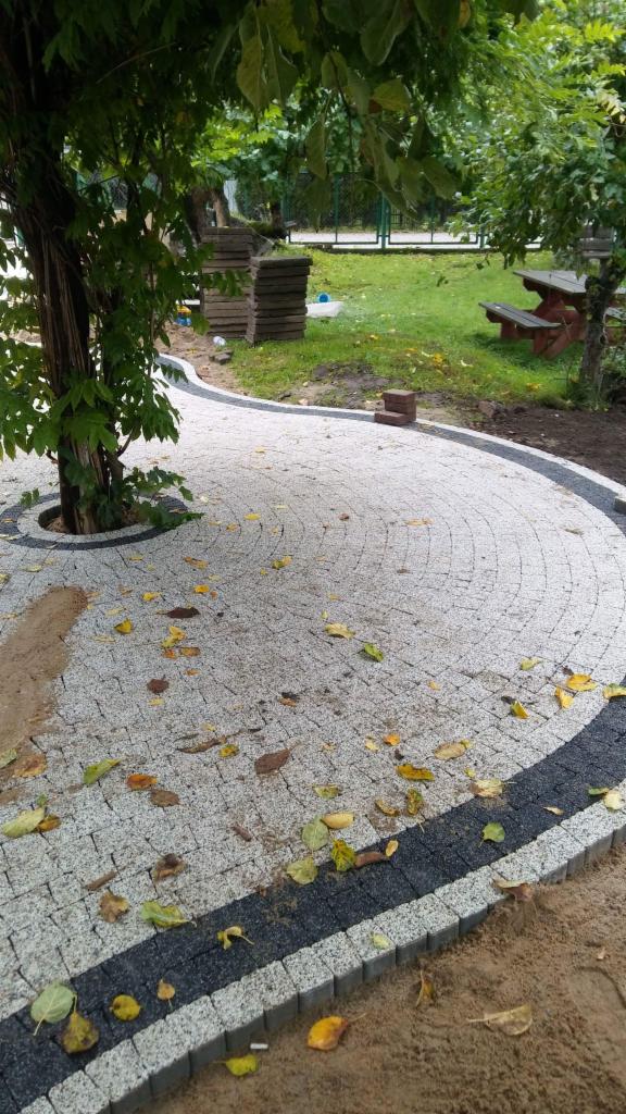 Ścieżka z jasnoszarej kostki brukowej ułożona wokół drzewa, z czarnym obrzeżem, na której leżą opadłe liście, w tle trawnik z ławką i stosem kostki.