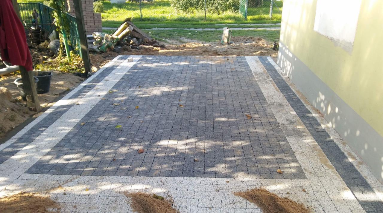 Ułożona szara kostka brukowa z białym obramowaniem, widoczne ślady piasku i cienie drzew, fragment ściany budynku w tle.
