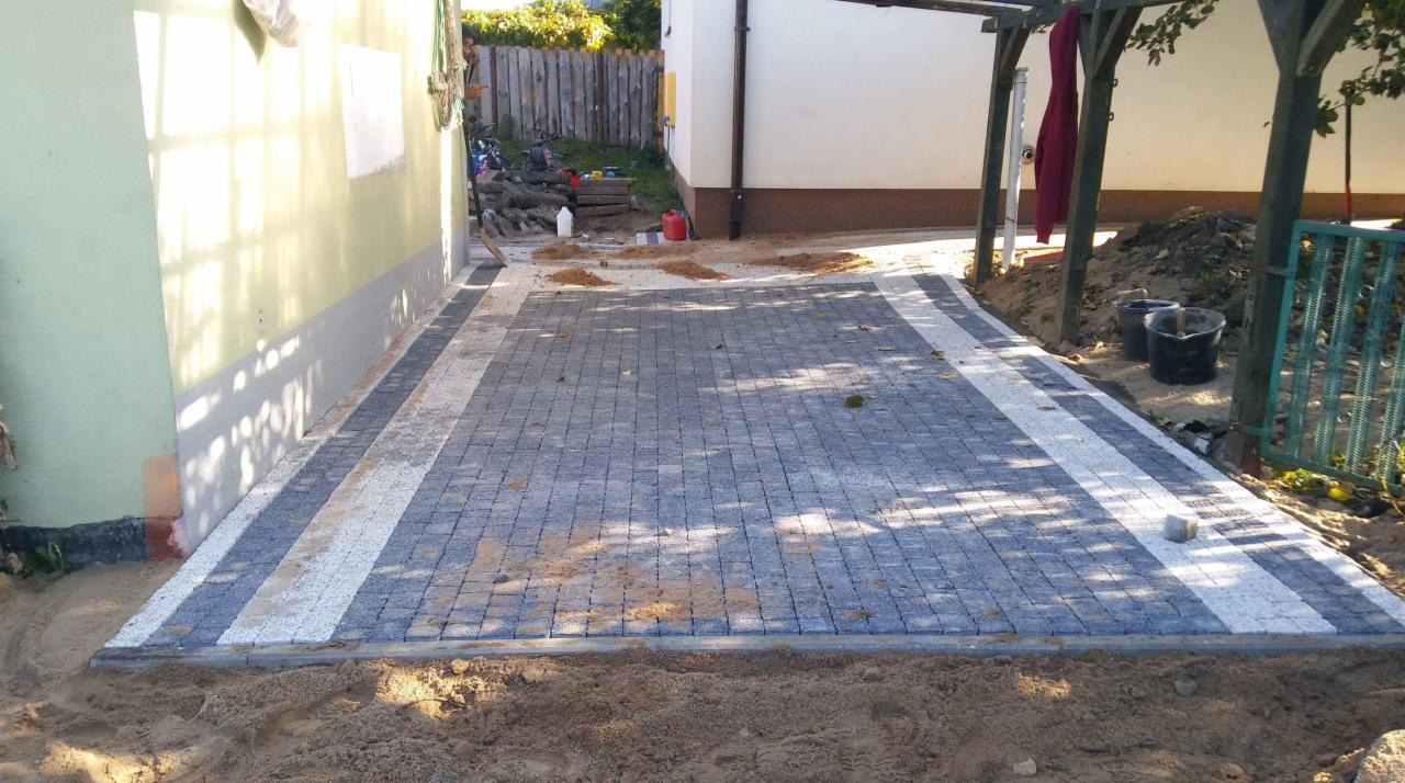 Ułożona szara kostka brukowa z białymi pasami obramowania, otoczona piaskiem, przylegająca do ściany budynku i drewnianej konstrukcji.