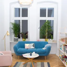 Jasne wnętrze gabinetu z błękitną sofą, różowym fotelem, regałem z książkami i roślinami doniczkowymi przy oknach.