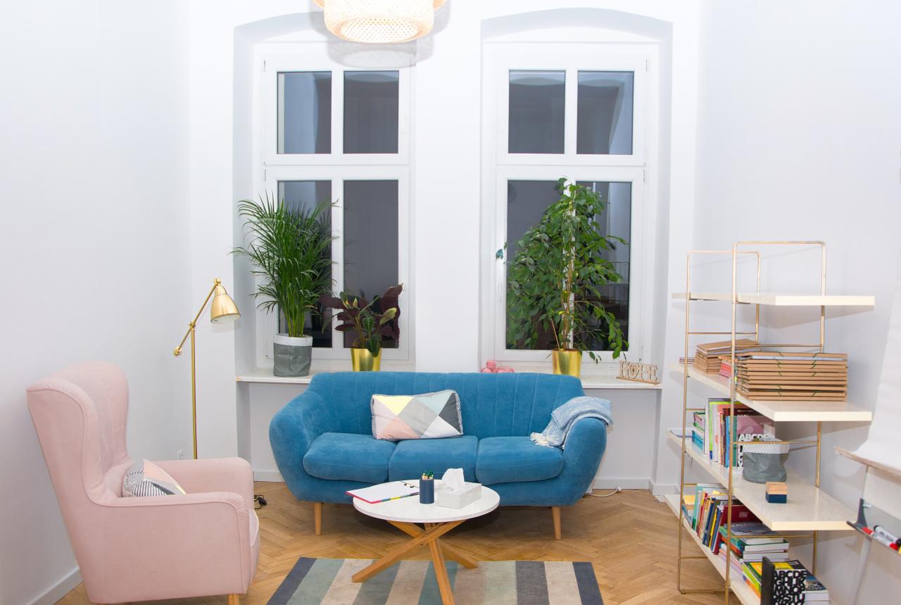 Jasne wnętrze gabinetu z błękitną sofą, różowym fotelem, regałem z książkami i roślinami doniczkowymi przy oknach.