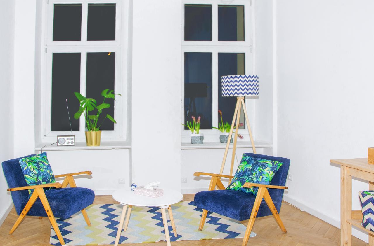 Jasny pokój z parą granatowych foteli z drewnianymi podłokietnikami, stolikiem kawowym, lampą na drewnianym trójnogu i roślinami w doniczkach na parapecie.