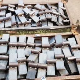 Metalowe zawiasy w skrzyni transportowej z widocznym napisem 'SCHAFER 0.5t/4.5t/', ułożone warstwami, gotowe do dalszej obróbki lub montażu.