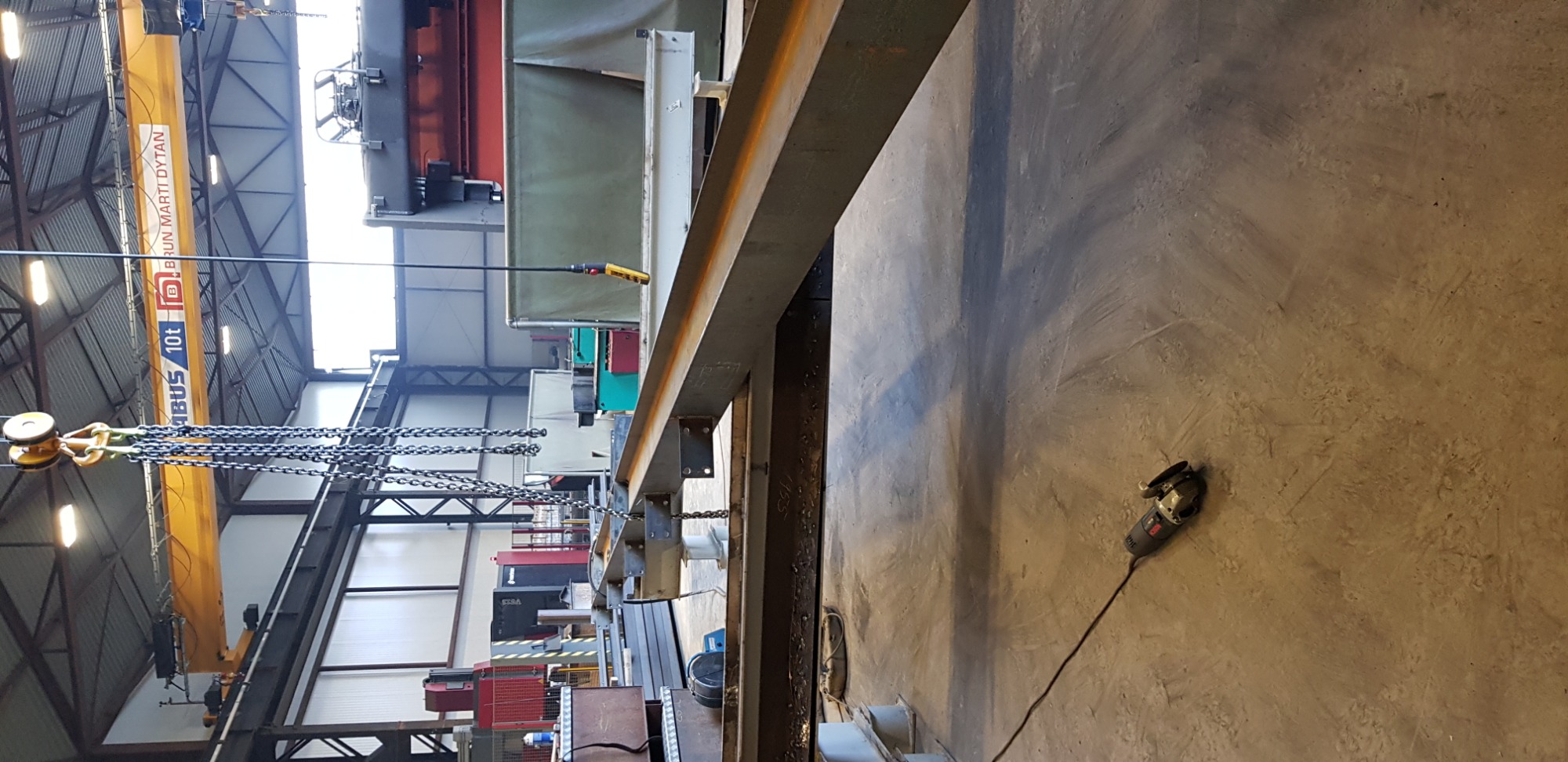 Wnętrze hali produkcyjnej z widoczną konstrukcją stalową, suwnicą z łańcuchami oraz szlifierką kątową leżącą na betonowej posadzce.