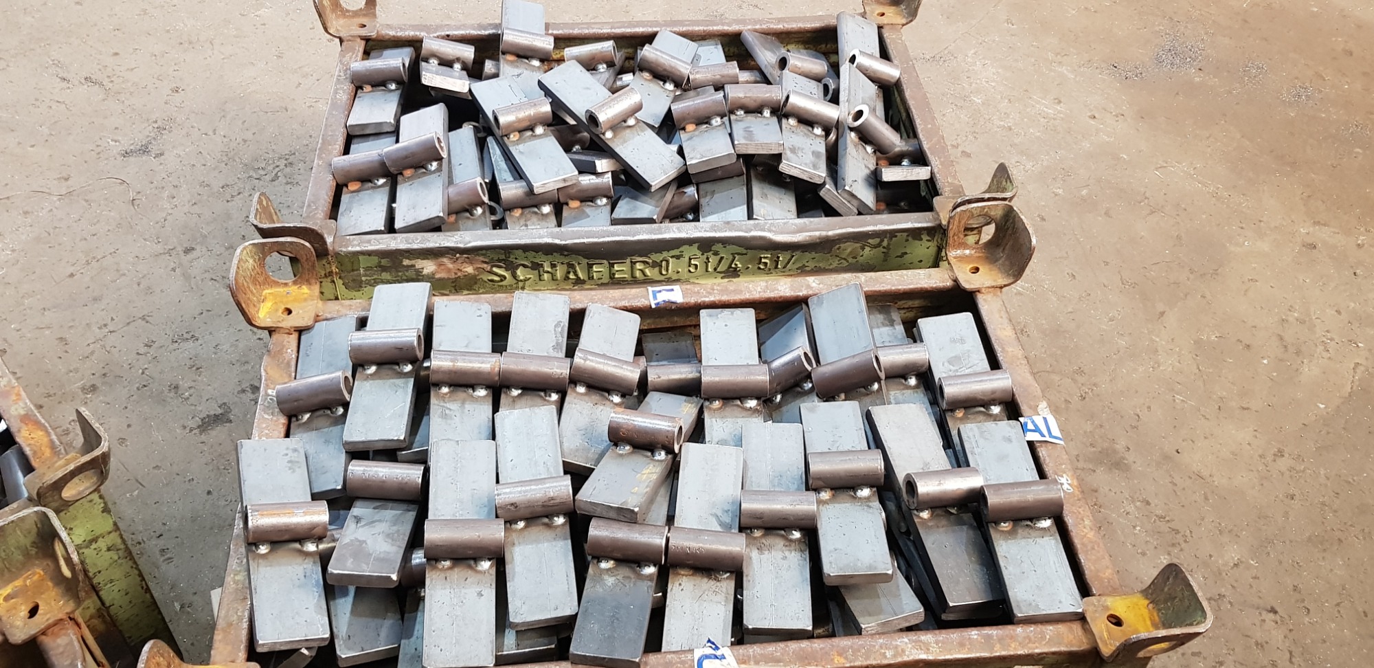 Metalowe zawiasy w skrzyni transportowej z widocznym napisem 'SCHAFER 0.5t/4.5t/', ułożone warstwami, gotowe do dalszej obróbki lub montażu.