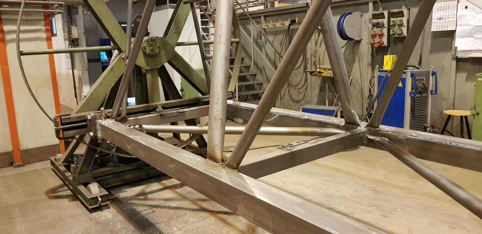 Surowa, metalowa konstrukcja szkieletowa z widocznymi spawami, ustawiona w hali produkcyjnej z zielono-szarą ścianą i schodami w tle.