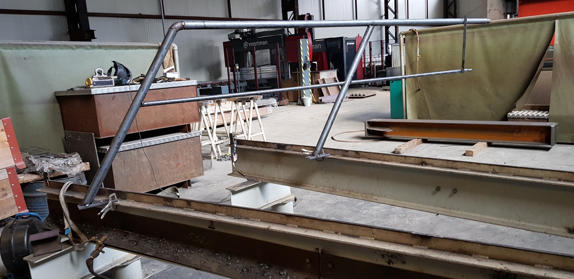 Metalowa konstrukcja z rur i profili na tle hali produkcyjnej, widoczne spawane łączenia i narzędzia pracy.