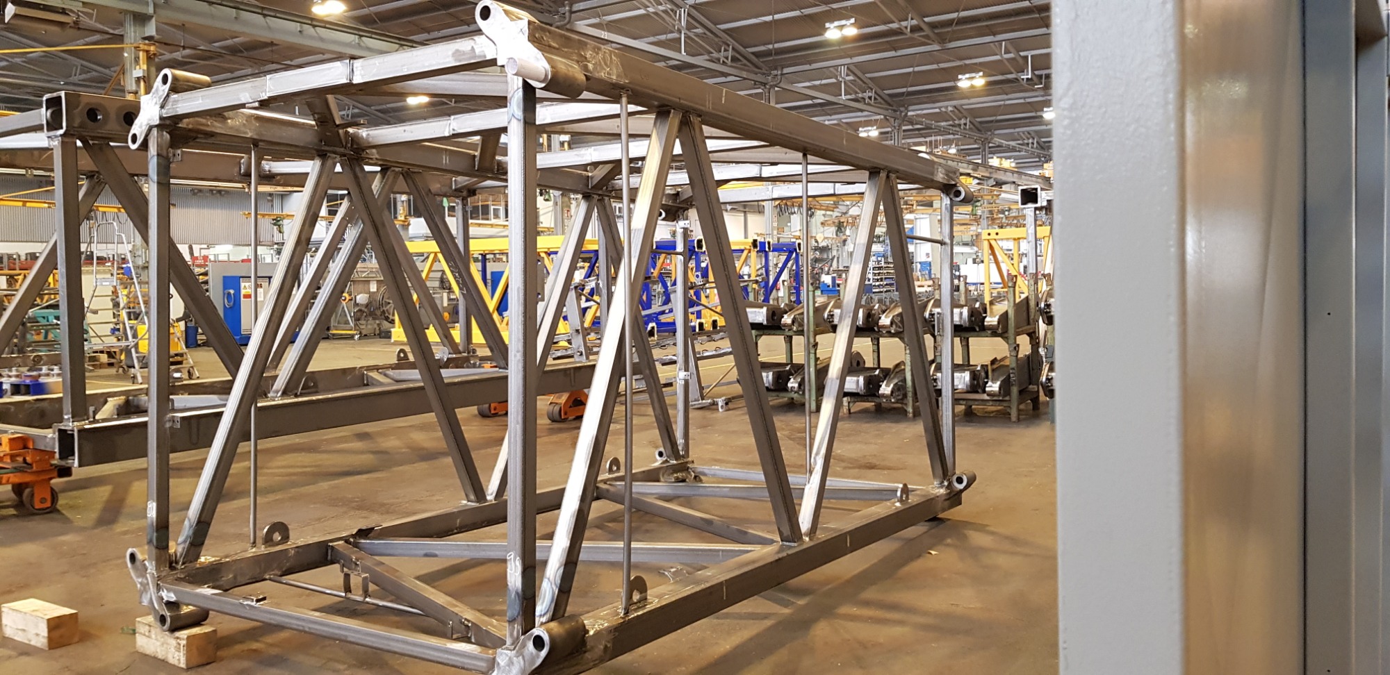 Surowa, metalowa konstrukcja hali w trakcie budowy, widoczna na tle hali produkcyjnej z wózkami i innymi konstrukcjami.