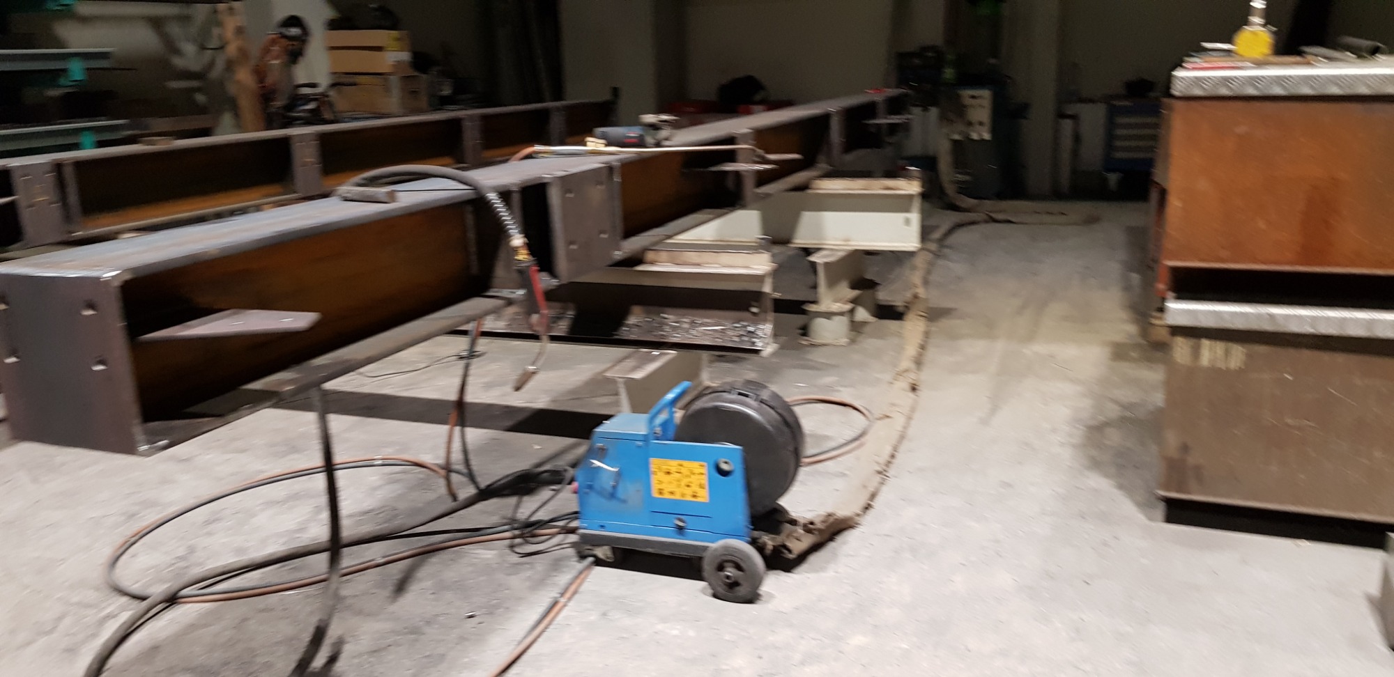 Wnętrze hali produkcyjnej z długimi, stalowymi belkami w trakcie montażu, widoczna spawarka z przewodami na podłodze.