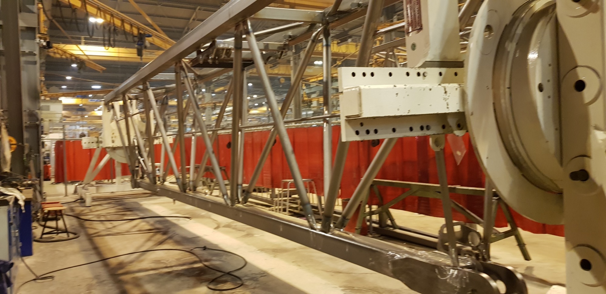 Wnętrze hali produkcyjnej z widoczną stalową konstrukcją kratownicową w trakcie budowy, z czerwonymi zasłonami spawalniczymi w tle i elementami mocującymi na końcach.