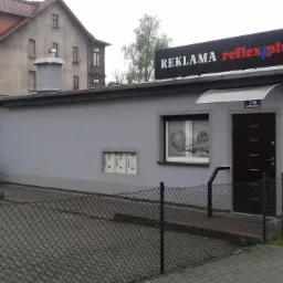 Budynek parterowy z szarymi ścianami, na dachu szyld z napisem 'REKLAMA reflex plus', okno z dekoracją, skrzynki pocztowe i brama wejściowa.