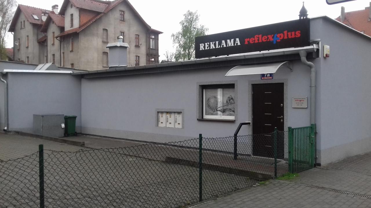 Budynek parterowy z szarymi ścianami, na dachu szyld z napisem 'REKLAMA reflex plus', okno z dekoracją, skrzynki pocztowe i brama wejściowa.