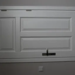 Biała, lakierowana klapa rewizyjna z metalowym uchwytem na tle szarej ściany.