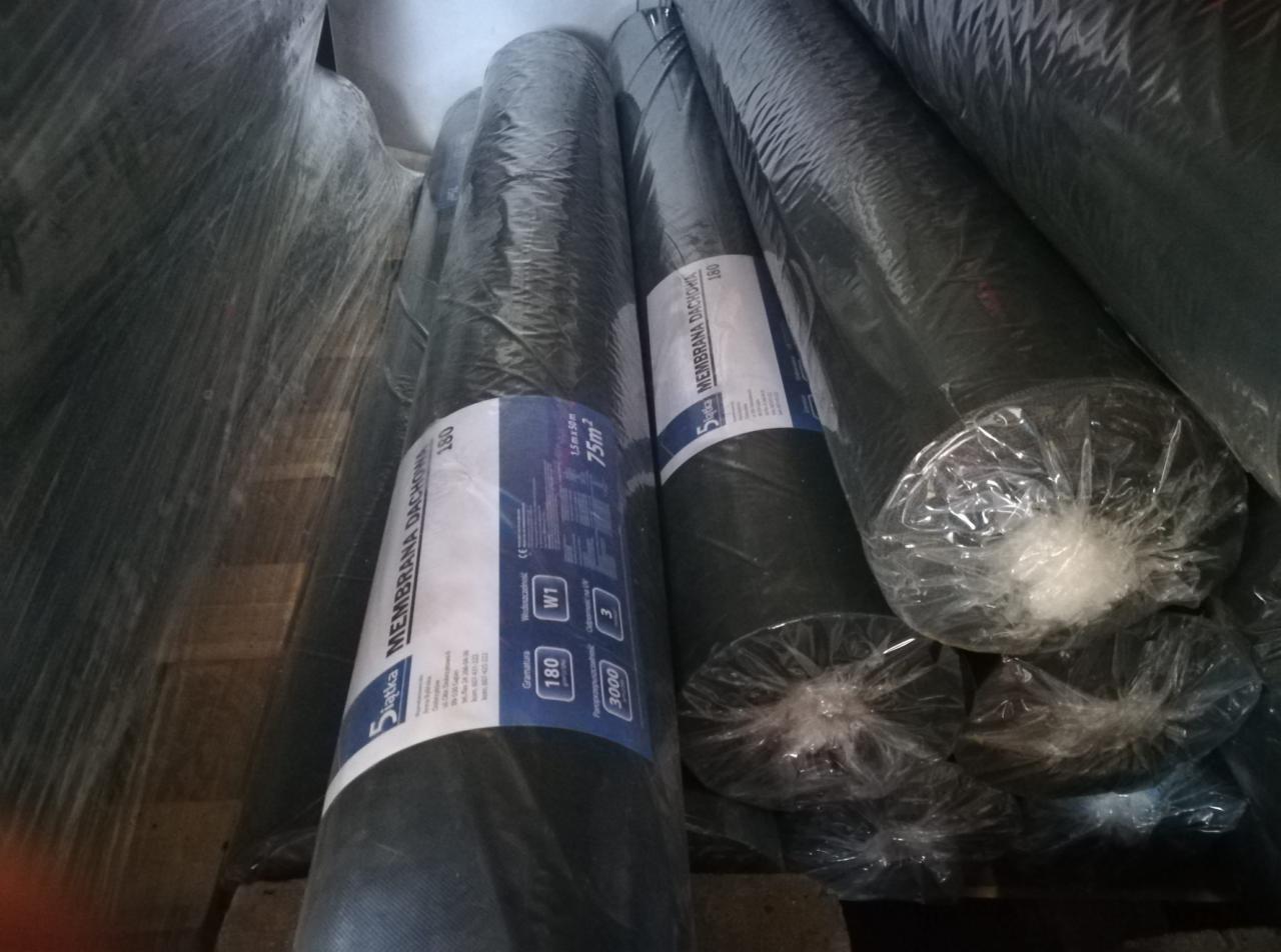 Stos rolek czarnej membrany dachowej, owiniętych folią, z widocznymi etykietami producenta Siatka, ułożonych na palecie w magazynie.