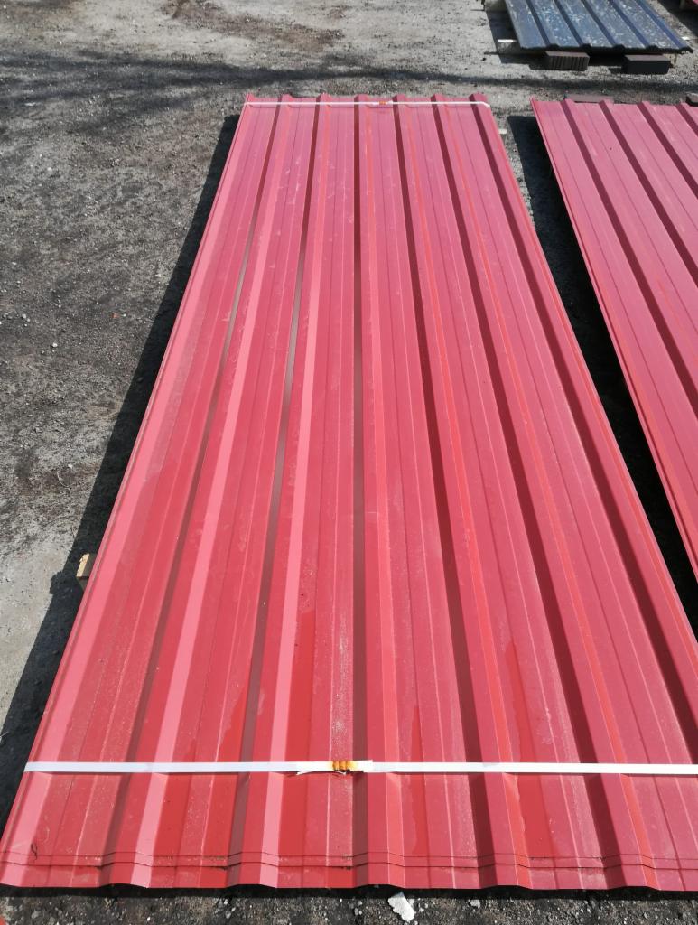 Czerwone blaszane panele dachowe, związane białą taśmą, leżące na ziemi. W tle widoczne ciemniejsze panele.