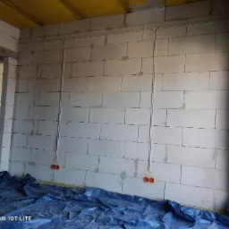 Wewnętrzna ściana z białych bloków z rozprowadzoną instalacją elektryczną w rurkach i pomarańczowymi puszkami pod gniazdka, zabezpieczona folią budowlaną na podłodze.