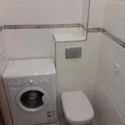 Wykończona łazienka z białą glazurą, dekoracyjnym paskiem z mozaiki, podwieszaną toaletą z chromowanym przyciskiem spłukiwania i pralką Indesit.