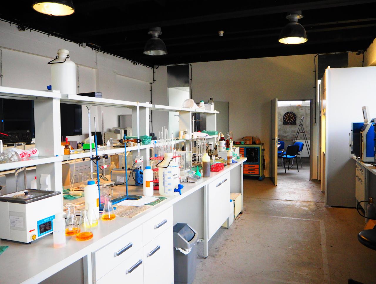 Wnętrze laboratorium z białymi meblami, szkłem laboratoryjnym, odczynnikami i sprzętem, widoczne lampy przemysłowe i otwarte drzwi do kolejnego pomieszczenia z tarczą do darta.