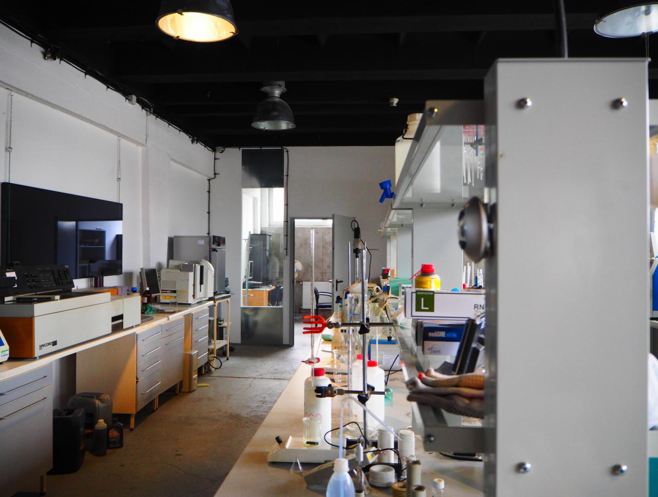Laboratoryjne wnętrze z białymi szafkami, stołami, szklanymi naczyniami i sprzętem pomiarowym, oświetlone lampami wiszącymi.