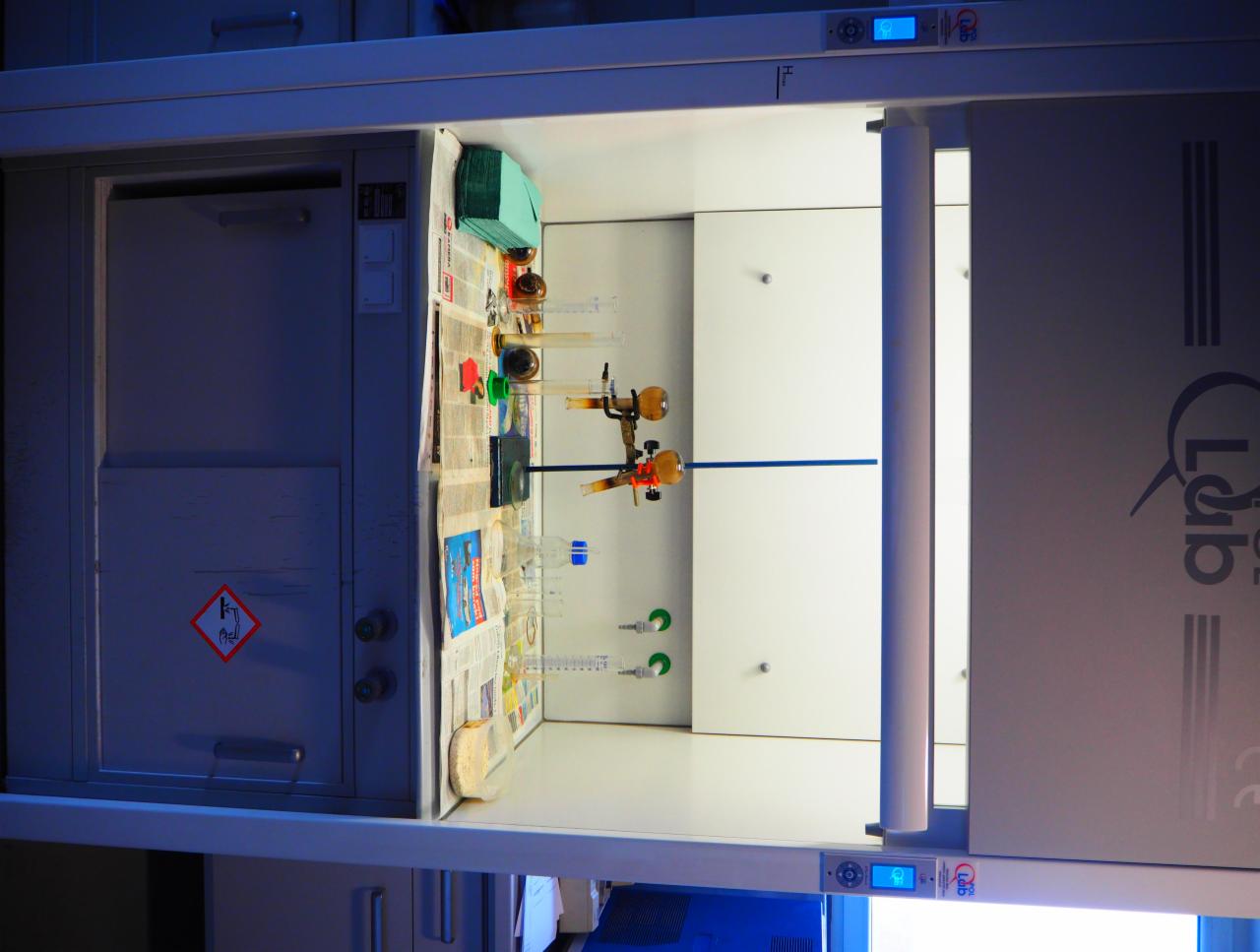 Wnętrze laboratoryjnej komory z aparaturą, szkłem laboratoryjnym i gazetą jako podkładem, widoczny znak ostrzegawczy na szafce po lewej stronie.