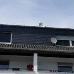 Dwupiętrowy dom z balkonem i dachem pokrytym ciemną dachówką, widoczne okna dachowe i dwie anteny satelitarne.