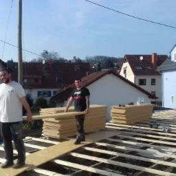 Dwóch mężczyzn układa płyty na drewnianej konstrukcji dachu, w tle zabudowa mieszkalna