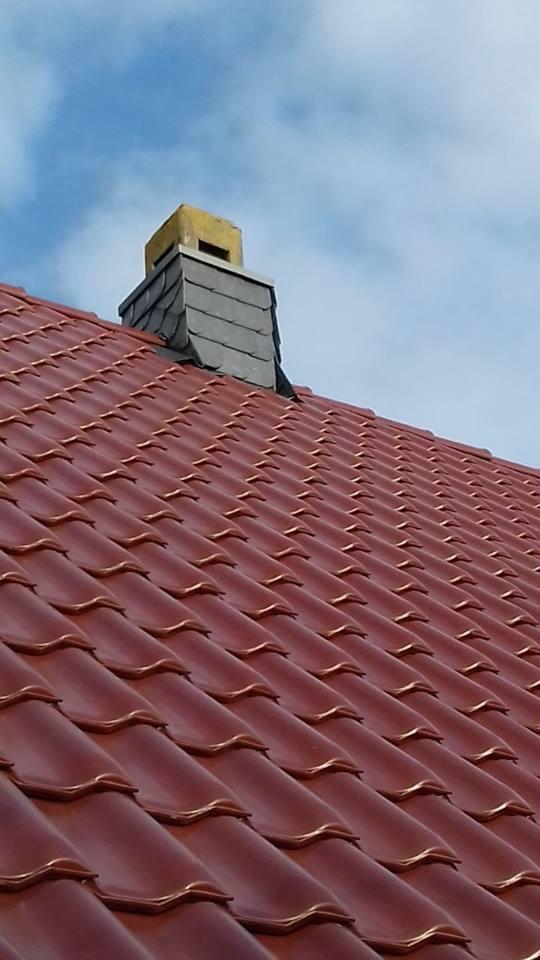 Czerwona dachówka ceramiczna na skośnym dachu z kominem wykończonym łupkiem i żółtym elementem, widok z dołu na tle błękitnego nieba.